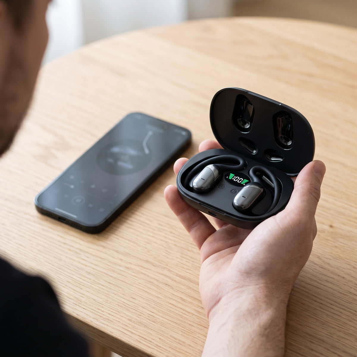 Main tenant l'étui de charge ouvert des écouteurs traducteurs à côté d'un smartphone pour montrer la taille réelle